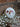 A single, round white bath bomb decorated with vibrant dried pink and orange flower petals, resting against a rustic brick wall with delicate white baby's breath flowers in the foreground.