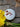 A high-angle, top-down shot of a single round, white bath bomb sprinkled with dried pink flower petals and orange rind, resting on a gray stone surface next to bright yellow flowers.
