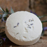 A single round, white bath bomb sprinkled with dried purple lavender buds, resting on a rustic stone surface with a sprig of fresh lavender in the background.