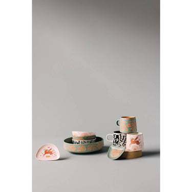 A collection of pink, teal, and black ceramic kitchenware featuring celestial and botanical illustrations, including a pink mug with a winged horse and a large teal bowl.