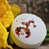 A single round, white bath bomb topped with vibrant dried rose and orange botanical petals, resting on a rustic stone surface next to a bright yellow flower.