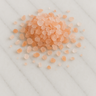 A loose mound of coarse pink Himalayan salt crystals, featuring various shades of peach and salmon pink, scattered on a white marble surface with light grey veining.