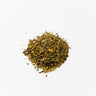 A small mound of dried holy basil leaves, featuring a textured mix of earthy green and brown fragments, cut and sifted on a clean white surface.