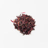 A small, centered mound of dried hibiscus flower petals, featuring a deep burgundy and crimson color with a slightly curled, leathery texture on a plain white surface.