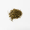 A small, centered pile of dried lemon balm leaves, consisting of finely crushed green and brown herbal fragments, staged on a clean white background.
