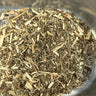 A high-angle, close-up view of dried meadowsweet herb, featuring a dense mixture of fragmented green leaves and pale tan, woody stem pieces inside a polished stainless steel container.