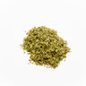 A small mound of dried yerba mate leaves, consisting of finely chopped herbal flakes in various shades of olive and forest green. The mixture is placed on a clean white background and shows a dry, slightly leathery texture.