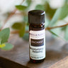An 8mL small bottle of 'Enchant' essential oil by Megan & Co. Apothecary, positioned on a wooden surface with green foliage in the background.