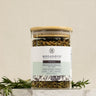 A glass jar containing loose leaf herbal tea blend with a label that reads 'FOCUS'. The jar is placed on a white surface with a hint of herbs visible around it.
