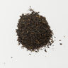 A small mound of loose-leaf orange pekoe black tea, consisting of fine, dark brown, slightly curled tea leaves with visible small golden-brown tip fragments, staged on a clean white background.