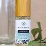 A glass spritzer bottle of Boji Bliss along with a glass bottle with measurement markings at the back and fresh green leaves on its base, staged on a clean white surface.