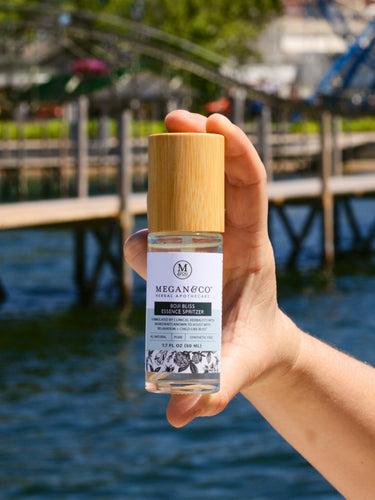 A hand holding the Megan & Co. Boji Bliss spritzer bottle with a wooden cap, staged against a scenic background featuring a Ferris wheel, a wooden bridge, and a glimpse of the sea.