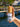 A hand holding the Megan & Co. Boji Bliss spritzer bottle with a wooden cap, staged against a scenic background featuring a Ferris wheel, a wooden bridge, and a glimpse of the sea.