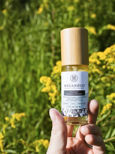 A hand holding a Megan & Co. Bouquet Essence Spritzer with a bamboo cap in a blurry grassy field with yellow wildflowers as its background.