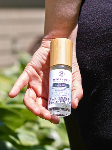 A hand holding a capped Megan & Co. Bug Ban spritzer bottle near a person's legs, featuring black pants and a lush, blurred green garden background.