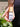 A hand holding a capped Megan & Co. Bug Ban spritzer bottle near a person's legs, featuring black pants and a lush, blurred green garden background.