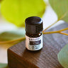 A 2.3ml amber glass bottle of Megan & Co. Green Clean essential oil on a wooden surface with a blurred leaf background, containing organic lemon, tea tree, and eucalyptus.