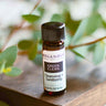 An 8ml amber glass bottle of Megan & Co. Green Clean essential oil on a wooden surface with a blurred leaf background, containing organic lemon, tea tree, and eucalyptus.