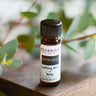 An 8ml amber glass vial of Megan & Co. Happiness essential oil on a wooden surface with a blurred leaf background, featuring uplifting notes of Lemon, Orange, and Jasmine.