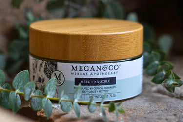 Megan & Co. Heel + Knuckle herbal salve jar with a bamboo lid on a rough stone surface with eucalyptus leaves on the side and a blurry background.