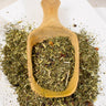 Mama + One loose leaf herbal tea on a wooden scoop, resting on scattered tea blend atop a white layered marble surface.