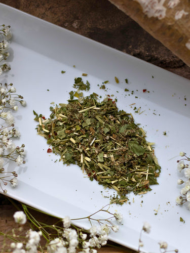 Mama + One loose leaf tea on a white plate with white flowers and a tree branch, featuring nourishing herbs like Nettle, Raspberry Leaf, and Oat Straw.
