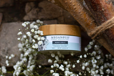 Megan & Co. Mama's Belly Herbal Whip jar with a bamboo lid on a rough surface, surrounded by delicate white flowers with rustic branches and rope overhead.