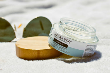 An open jar of Megan & Co. Mint Aloe body cream propped on its bamboo lid next to sunglasses on a white towel.