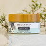 Megan & Co. Neroli Rose facial cream in a glass jar with a bamboo lid on a white marble surface with white waxflowers in the background.