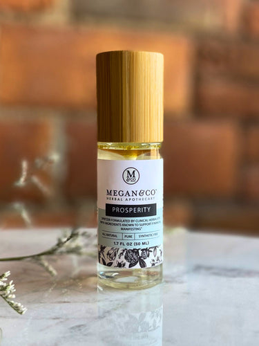 Prosperity room and body spritzer in a clear bottle with a wooden cap, staged on a white marble surface with tiny white flowers and a blurred rustic brick wall background.