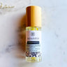 Megan & Co. Prosperity spritzer in a clear glass bottle with a wooden cap, laid on a white marble surface and staged with delicate white flowers.