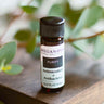 An 8ml amber glass bottle of Megan & Co. Purify essential oil on a wooden surface with a blurred leaf background, featuring Myrrh, Sage, and Rose Absolute.
