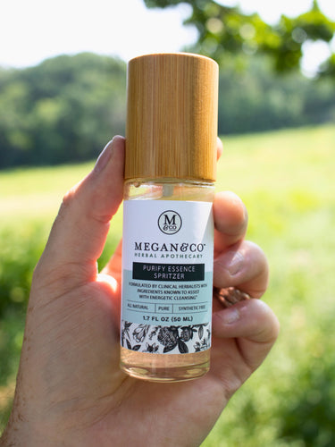 A hand holding the Purify room and body spritzer bottle with a wooden cap against a soft, blurry green grassy nature background.