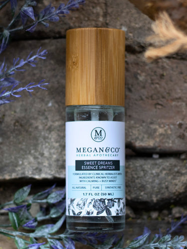 Megan & Co. Sweet Dreams Essence Spritzer with a bamboo cap on a rough stone surface with purple flowers and an overhead branch.
