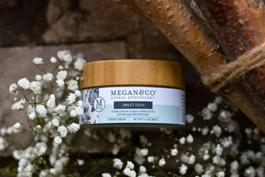 Sweet Tush herbal salve jar on a rough stone surface staged with baby's breath and twine-wrapped branches.