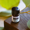 A 2.3ml amber glass bottle of Megan & Co. Winter Warmer essential oil on a wooden surface with a blurred leaf background, featuring grounding notes of Fir Balsam, Cedarwood, Orange, Clove, and Rosemary.