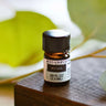 A 2.3ml (5/8 dram) amber glass vial of Megan & Co. Woods essential oil on a wooden surface with a blurred leaf background, featuring a grounding blend of Organic Fir Balsam, Spruce, and Cedarwood.