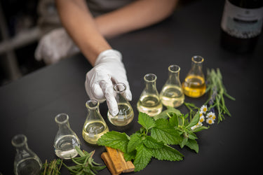 Person wearing gloves with various glass bottles and herbs on a dark surface