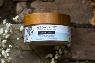 Megan & Co. Neroli Rose facial cream in a glass jar with a bamboo lid on a rough stone surface, staged with delicate white baby's breath flowers.