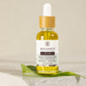 A small clear glass bottle with a dropper filled with yellow liquid, labeled 'Megan and Co R + R Herbal Oil', displayed on a white textured surface with a green leaf beside it.