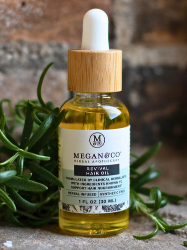 Bottle of Megan & Co. Revival Hair Oil with a wooden dropper on a stone surface with greenery.