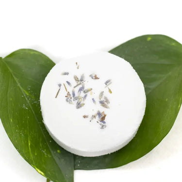 A direct overhead shot of a circular white bath bomb centered on two vibrant green leaves, showcasing a delicate cluster of dried purple lavender buds on its surface.