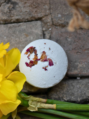A high-angle, top-down shot of a single round, white bath bomb sprinkled with dried pink flower petals and orange rind, resting on a gray stone surface next to bright yellow flowers.
