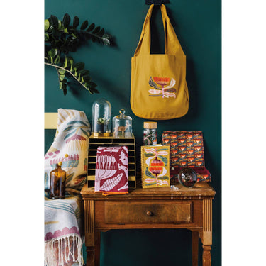 A vibrant lifestyle display of the Venus Sustain collection featuring the botanical glass tea tumbler on a wooden nightstand alongside a mustard yellow tote bag, a floral notebook, and decorative textiles. The scene is set against a deep teal wall and includes small glass cloches and a fringed throw blanket with matching floral patterns.