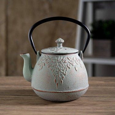 A small cast iron teapot with a textured light green and beige finish, featuring embossed floral patterns, a black metal handle, and a silver knob on the lid, set on a wooden table.