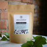 A 1oz sample bag of Megan & Co. Hope + Love tea on a white marble table, staged with green leaves at the base and a blurred brick wall in the background.